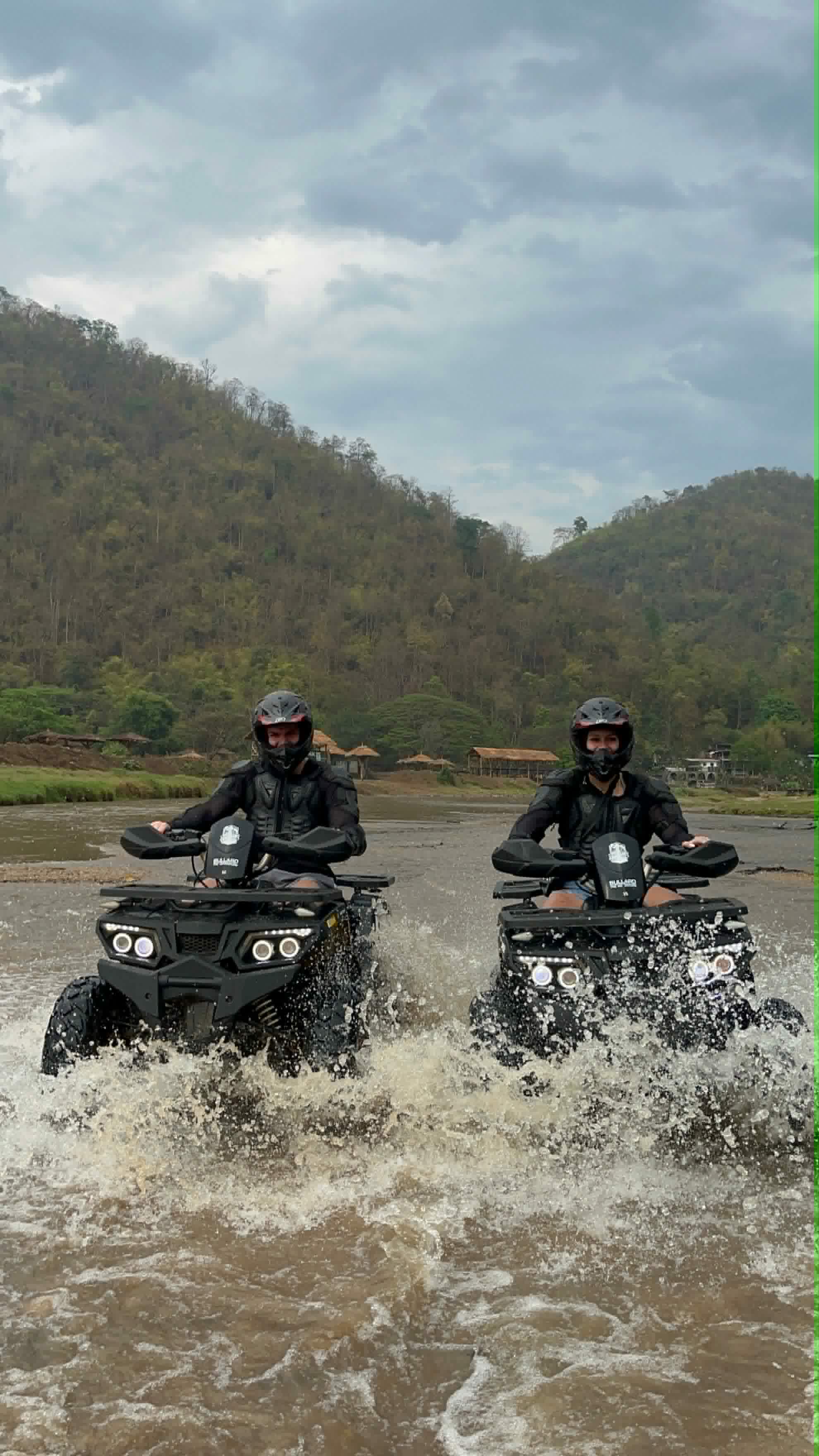Two guests riding ATVs through a river splash in Mae Taeng
